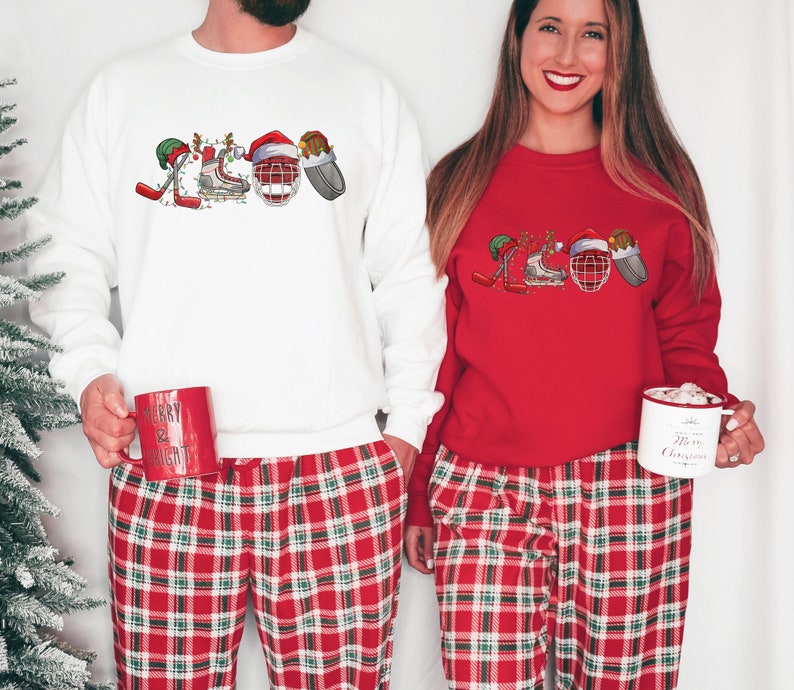 May include: A couple wearing matching red and white plaid pajamas with Christmas-themed hockey player graphics on their white and red sweatshirts. The man is holding a red mug that says "Merry & Bright" and the woman is holding a white mug that says "It's the Most Wonderful Time of the Year".