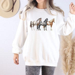 May include: White sweatshirt with a graphic of three horses in a row. The horses are brown, black, and white.