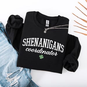 May include: Black sweatshirt with white text that reads "Shenanigans coordinator" and a green four-leaf clover.