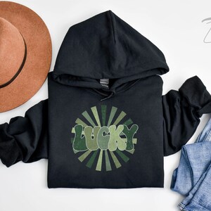 Peut inclure: Sweat-shirt &agrave; capuche noir avec un motif vert et blanc qui dit "LUCKY" dans une police r&eacute;tro. Le motif est entour&eacute; d'un design en forme de soleil.