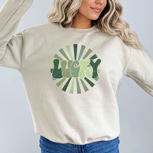 Peut inclure: Un sweat-shirt beige clair avec un motif r&eacute;tro vert et blanc qui dit "LUCKY" dans un design en forme de soleil.