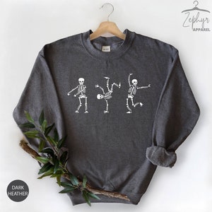 May include: Dark heather grey sweatshirt with a white graphic of three skeletons dancing. The skeletons are in different poses, with one skeleton doing a backflip.