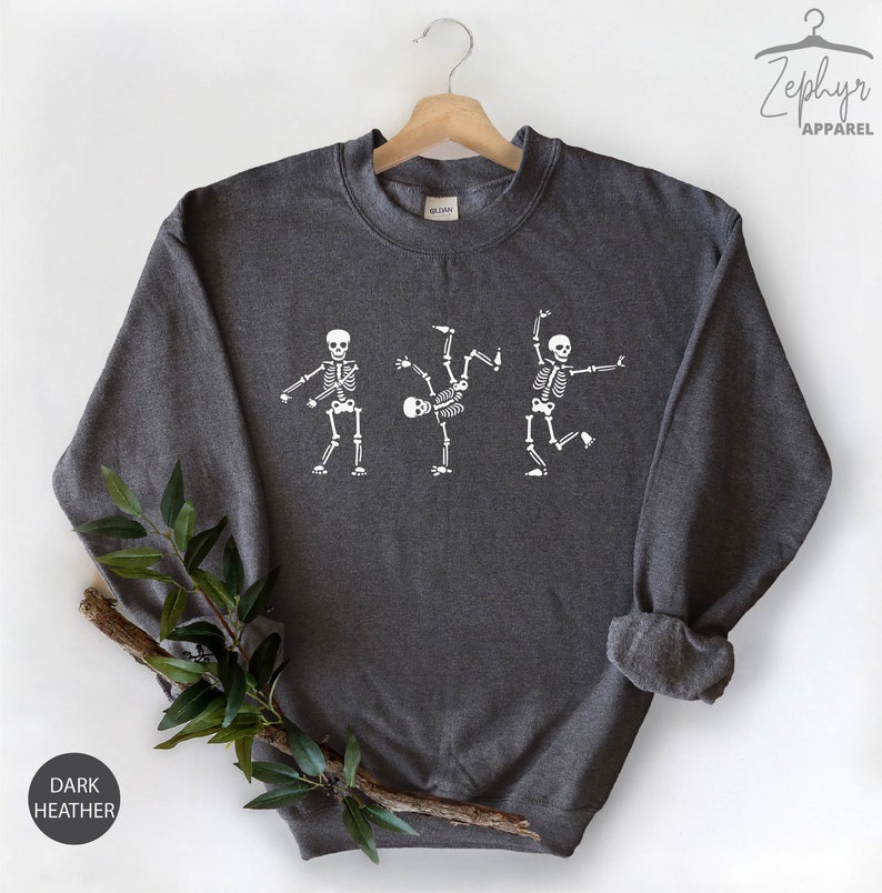 May include: Dark heather grey sweatshirt with a white graphic of three skeletons dancing. The skeletons are in different poses, with one skeleton doing a backflip.