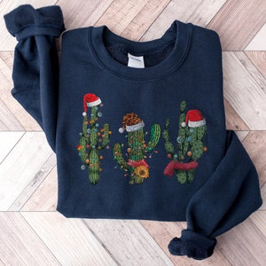 May include: A navy blue sweatshirt with a Christmas cactus design. The cactus is wearing a Santa hat and has lights around it.