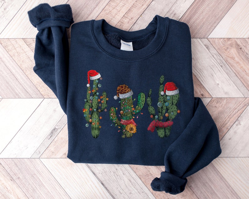 May include: A navy blue sweatshirt with a Christmas cactus design. The cactus is wearing a Santa hat and has lights around it.