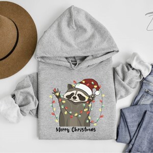 May include: A gray hooded sweatshirt with a cute cartoon raccoon wearing a Santa hat and holding Christmas lights. The text "Merry Christmas" is printed below the raccoon.