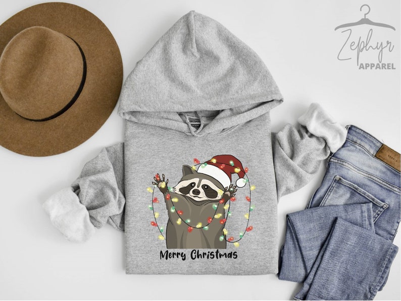 May include: A gray hooded sweatshirt with a cute cartoon raccoon wearing a Santa hat and holding Christmas lights. The text "Merry Christmas" is printed below the raccoon.