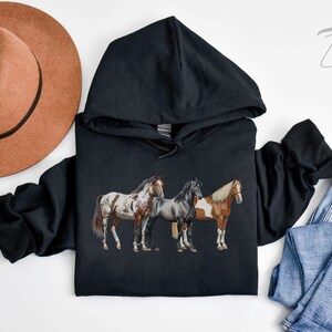 May include: Black hooded sweatshirt with a graphic of three horses. The horses are brown, white, and black with brown, white, and black spots. The sweatshirt has a hood with a drawstring.