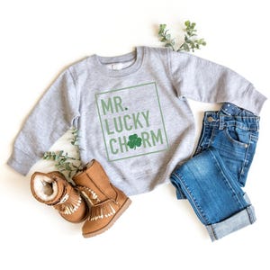 May include: A gray sweatshirt with the text "MR. LUCKY CHARM" in a green box. The sweatshirt is paired with blue jeans and brown boots.