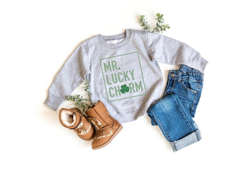 May include: A gray sweatshirt with the text "MR. LUCKY CHARM" in a green box. The sweatshirt is paired with blue jeans and brown boots.