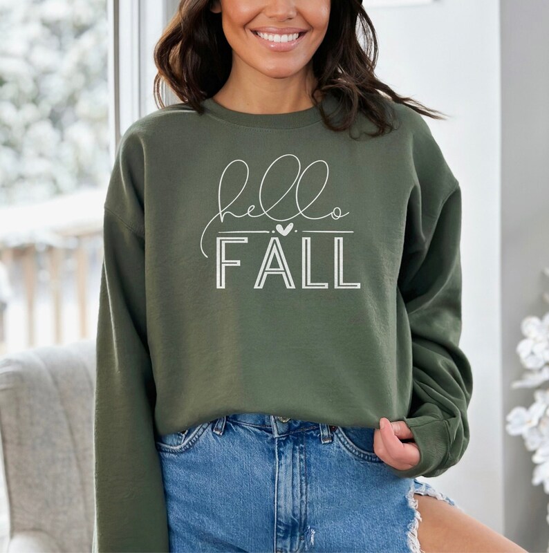 Op de afbeelding: Een groen sweatshirt met de tekst "hello fall" in witte letters. De tekst bevat een klein hartje.