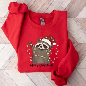 May include: Red Christmas sweatshirt with a raccoon wearing a Santa hat and tangled in Christmas lights. The text "Merry Christmas" is printed below the raccoon.