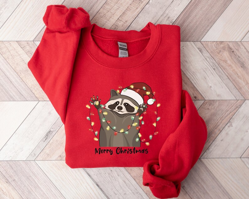 May include: Red Christmas sweatshirt with a raccoon wearing a Santa hat and tangled in Christmas lights. The text "Merry Christmas" is printed below the raccoon.