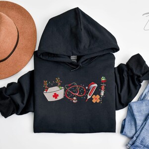 Puede incluir: Sudadera con capucha negra con un dise&ntilde;o navide&ntilde;o que incluye un gorro de enfermera, un estetoscopio, un term&oacute;metro, una venda y una jeringa. El dise&ntilde;o est&aacute; rodeado de luces navide&ntilde;as.