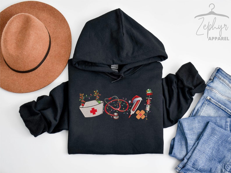 Puede incluir: Sudadera con capucha negra con un dise&ntilde;o navide&ntilde;o que incluye un gorro de enfermera, un estetoscopio, un term&oacute;metro, una venda y una jeringa. El dise&ntilde;o est&aacute; rodeado de luces navide&ntilde;as.