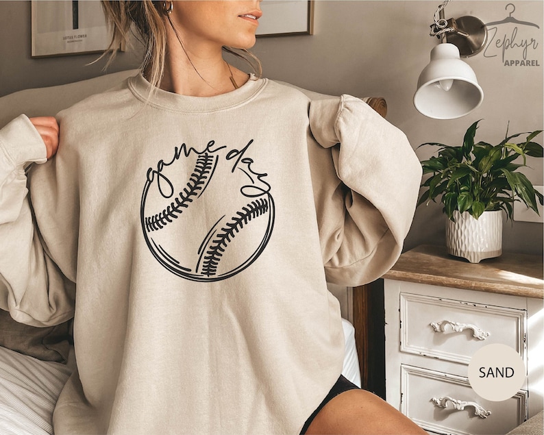 May include: A beige sweatshirt with a black baseball graphic and the text "game day" on the front.