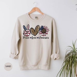 May include: A beige sweatshirt with a peace sign, heart, and sunflower design. The peace sign is red, white, and blue, the heart is leopard print, and the sunflower is yellow with a red center. The text "Peace Love America" is printed below the design.