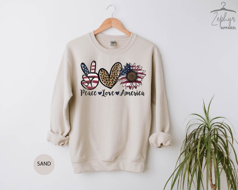 May include: A beige sweatshirt with a peace sign, heart, and sunflower design. The peace sign is red, white, and blue, the heart is leopard print, and the sunflower is yellow with a red center. The text "Peace Love America" is printed below the design.