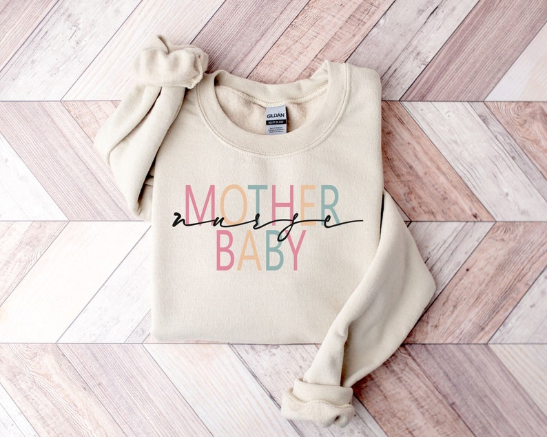 Puede incluir: Una sudadera beige con el texto "MOTHER" en naranja, "nurse" en rosa y "BABY" en azul y verde.