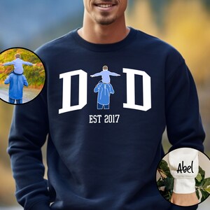 May include: Navy blue sweatshirt with white text that reads "DAD EST 2017". The sweatshirt features a graphic of a father carrying a child on his shoulders. The sleeve of the sweatshirt has white text that reads "Abel".
