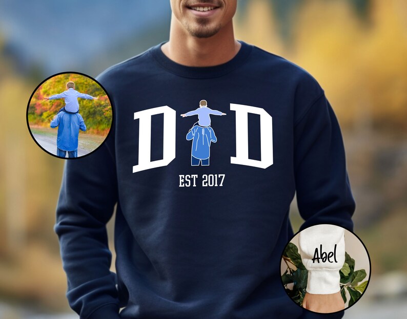 May include: Navy blue sweatshirt with white text that reads "DAD EST 2017". The sweatshirt features a graphic of a father carrying a child on his shoulders. The sleeve of the sweatshirt has white text that reads "Abel".