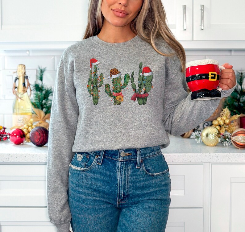 May include: A gray sweatshirt with a festive design of four cacti wearing Santa hats and decorated with Christmas lights.