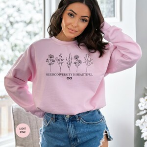 Op de afbeelding: Een lichtroze sweatshirt met een zwarte afbeelding van bloemen en de tekst "Neurodiversity is Beautiful" met een oneindigheidssymbool.