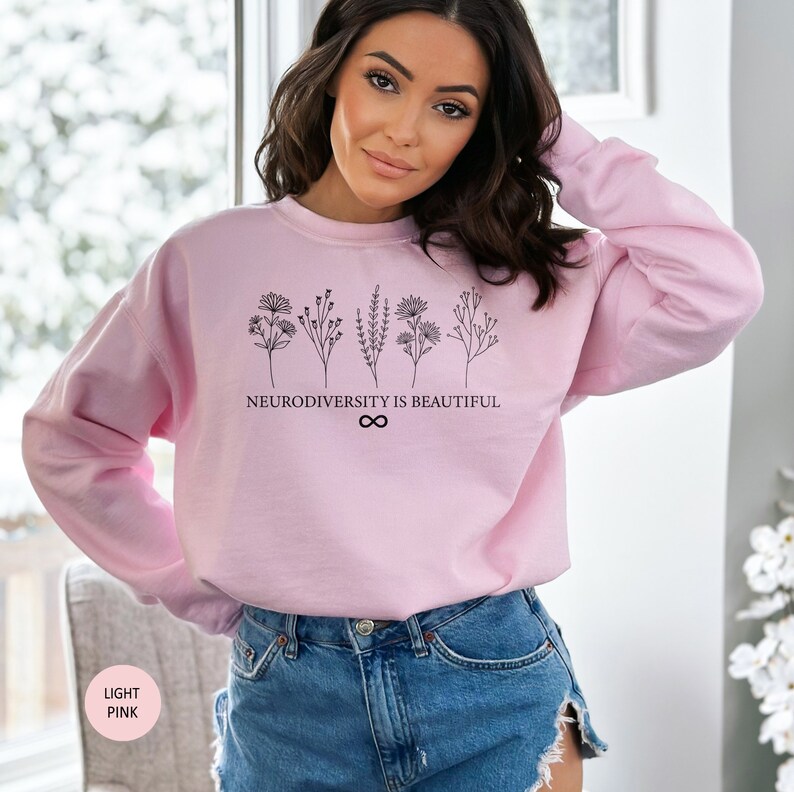 Op de afbeelding: Een lichtroze sweatshirt met een zwarte afbeelding van bloemen en de tekst "Neurodiversity is Beautiful" met een oneindigheidssymbool.