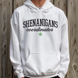 May include: White hooded sweatshirt with the text "Shenanigans coordinator" in black lettering and a green four-leaf clover.