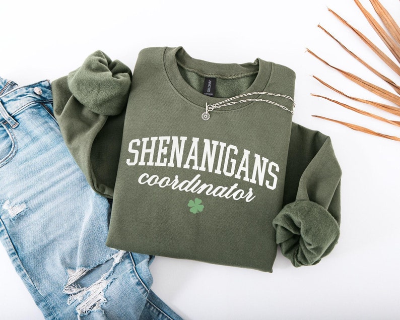 May include: A green sweatshirt with the text "SHENANIGANS coordinator" in white lettering and a green four-leaf clover graphic.