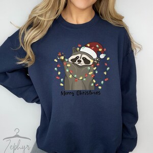 May include: A navy blue sweatshirt with a cartoon raccoon wearing a Santa hat and holding Christmas lights. The text "Merry Christmas" is printed below the raccoon.