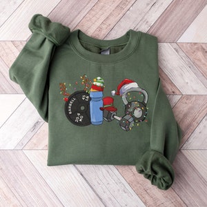 K&ouml;nnte beinhalten: Ein gr&uuml;nes Sweatshirt mit einem weihnachtlichen Design, das eine Langhantel, eine Kettlebell, eine Wasserflasche und eine Gewichtsplatte zeigt. Das Design enth&auml;lt Weihnachtslichter und Weihnachtsm&uuml;tzen.