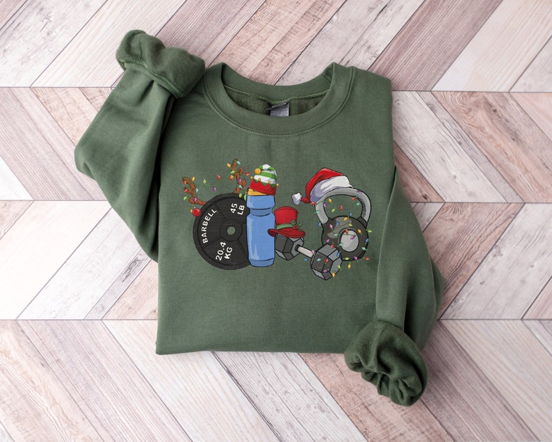 K&ouml;nnte beinhalten: Ein gr&uuml;nes Sweatshirt mit einem weihnachtlichen Design, das eine Langhantel, eine Kettlebell, eine Wasserflasche und eine Gewichtsplatte zeigt. Das Design enth&auml;lt Weihnachtslichter und Weihnachtsm&uuml;tzen.