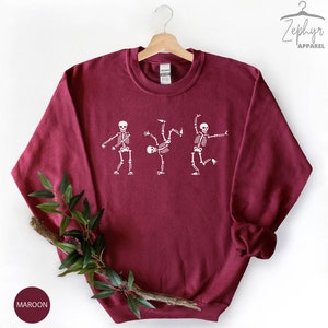 May include: Maroon sweatshirt with a white graphic of three skeletons dancing. The skeletons are in different poses, with one doing a handstand. The sweatshirt has a crew neck and long sleeves.