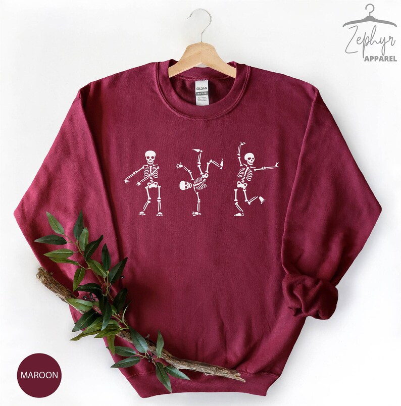 May include: Maroon sweatshirt with a white graphic of three skeletons dancing. The skeletons are in different poses, with one doing a handstand. The sweatshirt has a crew neck and long sleeves.