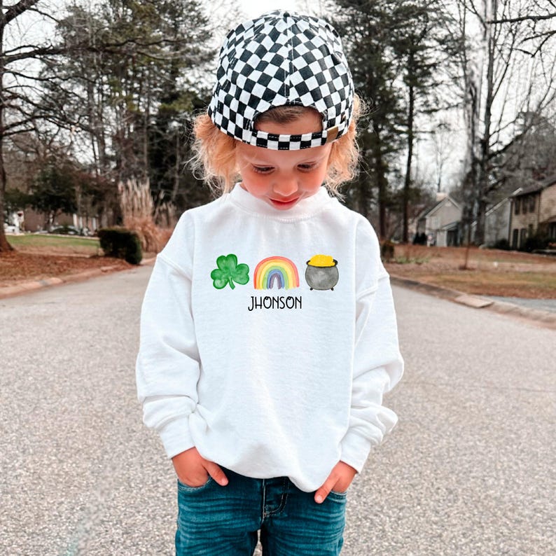 May include: A white sweatshirt with a green shamrock, rainbow, and pot of gold graphic. The text "JHONSON" is printed below the graphic. A child is wearing the sweatshirt and a black and white checkered hat.