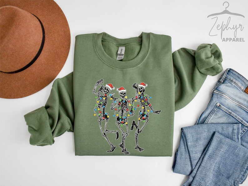 May include: A green sweatshirt with a festive design of three skeletons wearing Santa hats and Christmas lights. The skeletons are dancing and smiling.