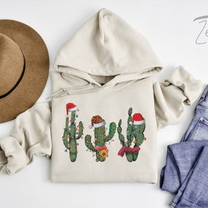 May include: A beige hoodie with a graphic of three cacti wearing Santa hats and Christmas lights. The cacti are green and have red and white Santa hats. The hoodie is on a white surface with a brown hat and blue jeans.