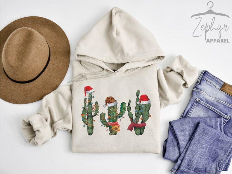 May include: A beige hoodie with a graphic of three cacti wearing Santa hats and Christmas lights. The cacti are green and have red and white Santa hats. The hoodie is on a white surface with a brown hat and blue jeans.