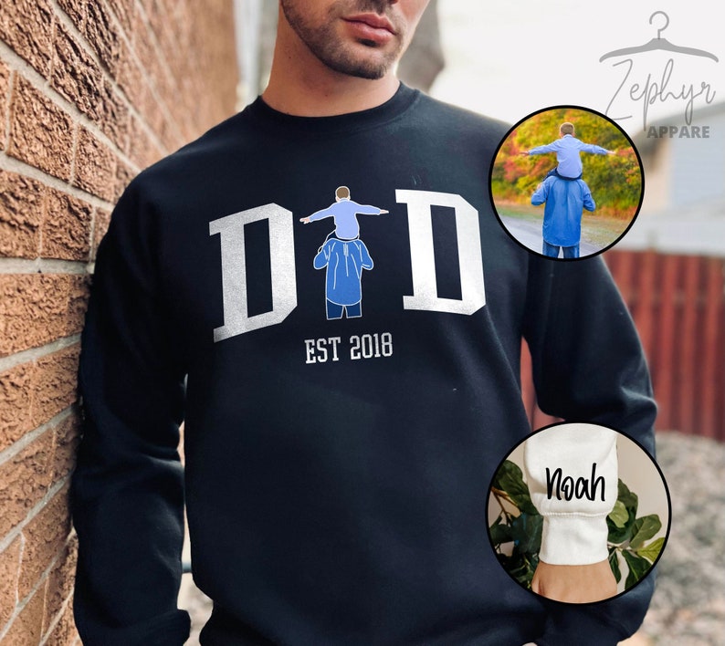 May include: Black sweatshirt with white text that reads "DAD EST 2018" and a graphic of a father carrying a child on his shoulders. The sweatshirt has a white cuff with the name "Noah" written in black.