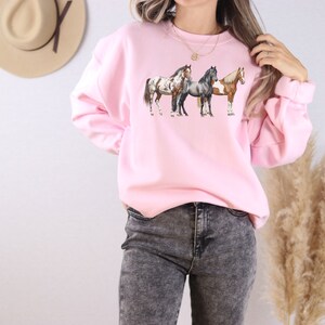 May include: A pink sweatshirt with a graphic of three horses in a line. The horses are brown, white, and gray.