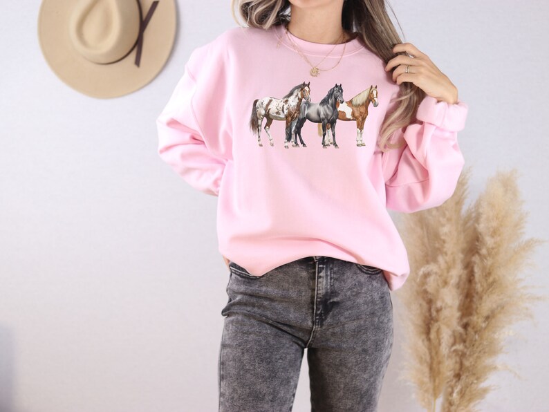 May include: A pink sweatshirt with a graphic of three horses in a line. The horses are brown, white, and gray.