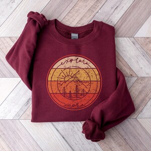 May include: Burgundy sweatshirt with a vintage-style graphic of a mountain range, sun, and trees. The graphic is in shades of orange, yellow, and red. The text "explore more" is written in a circle around the graphic.