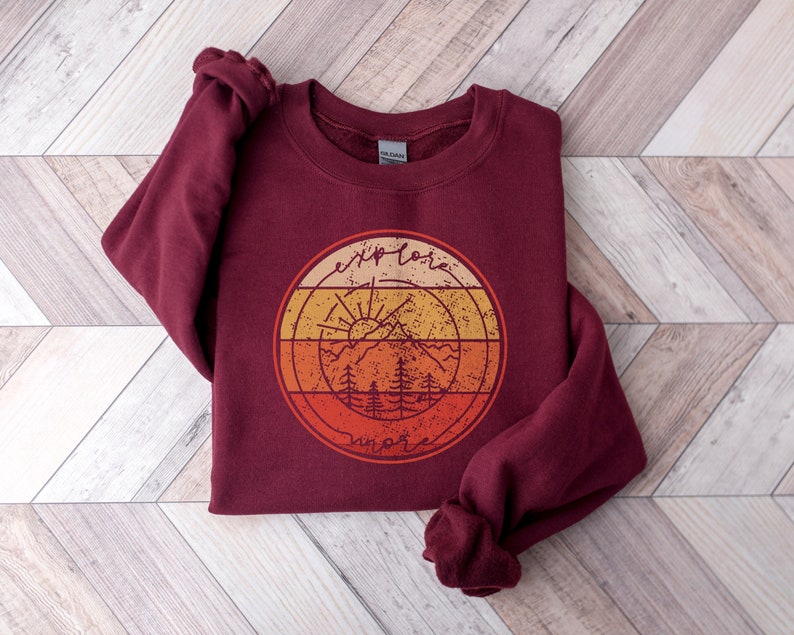 May include: Burgundy sweatshirt with a vintage-style graphic of a mountain range, sun, and trees. The graphic is in shades of orange, yellow, and red. The text "explore more" is written in a circle around the graphic.