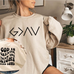 以下が含まれることがあります： 前面に黒い文字で「G>A」、背面に黒い文字で「God is greater than the highs and lows」と書かれたベージュのトレーナー。トレーナーは人が着用しています。