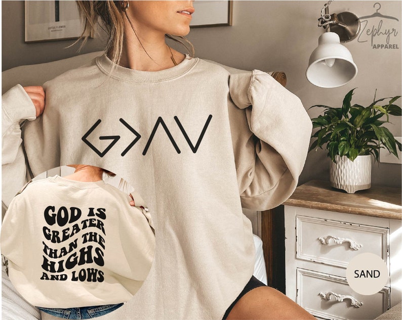 以下が含まれることがあります： 前面に黒い文字で「G>A」、背面に黒い文字で「God is greater than the highs and lows」と書かれたベージュのトレーナー。トレーナーは人が着用しています。