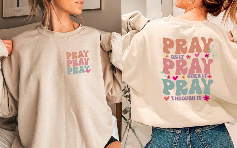 May include: A beige sweatshirt with a graphic print of the word "PRAY" repeated three times in different colours and styles. The sweatshirt has a crew neck and long sleeves.
