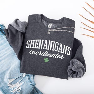 May include: A dark gray sweatshirt with the text "Shenanigans Coordinator" in white lettering and a green shamrock. The sweatshirt has a ribbed crew neck and long sleeves.