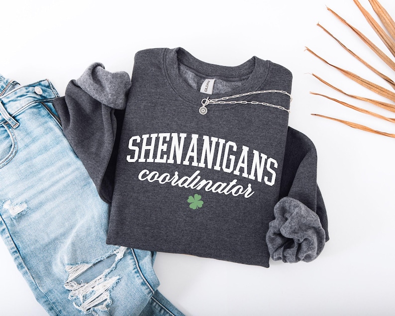 May include: A dark gray sweatshirt with the text "Shenanigans Coordinator" in white lettering and a green shamrock. The sweatshirt has a ribbed crew neck and long sleeves.