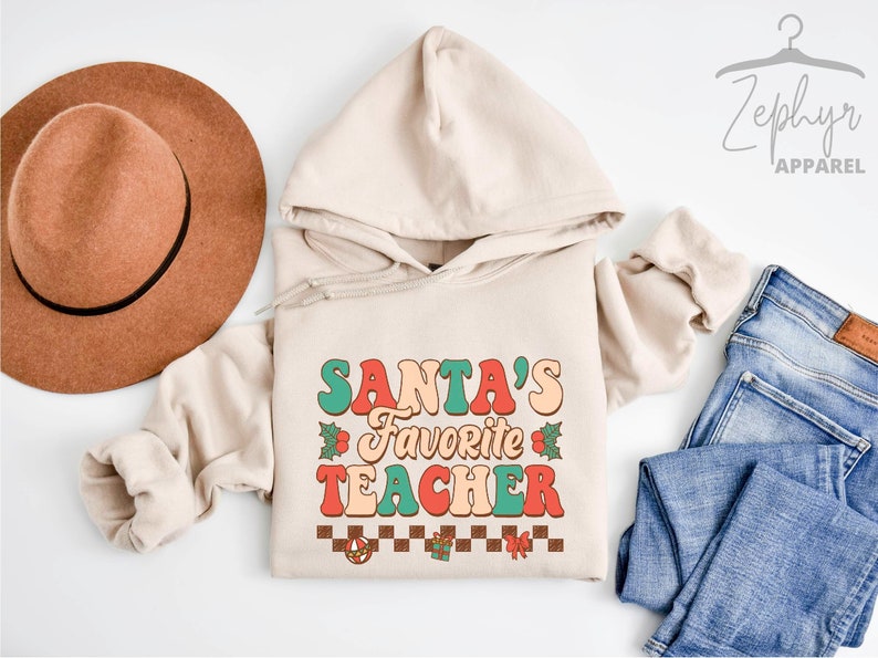 K&ouml;nnte beinhalten: Ein beiger Hoodie mit dem Text "SANTA'S FAVORITE TEACHER" in Retro-Schrift. Der Text ist in Rot, Gr&uuml;n und Orange mit einem karierten Muster und weihnachtlichen Grafiken darunter.